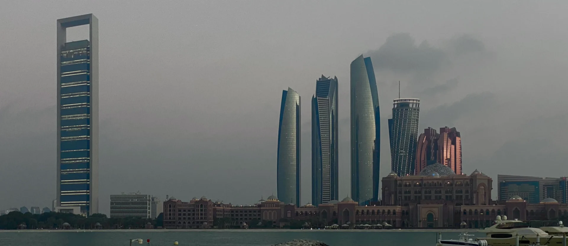 The Abu Dhabi skyline seen across the water under a grey sky: the ADNOC headquarters tower on the left, the Etihad Towers and the Emirates Palace on the right