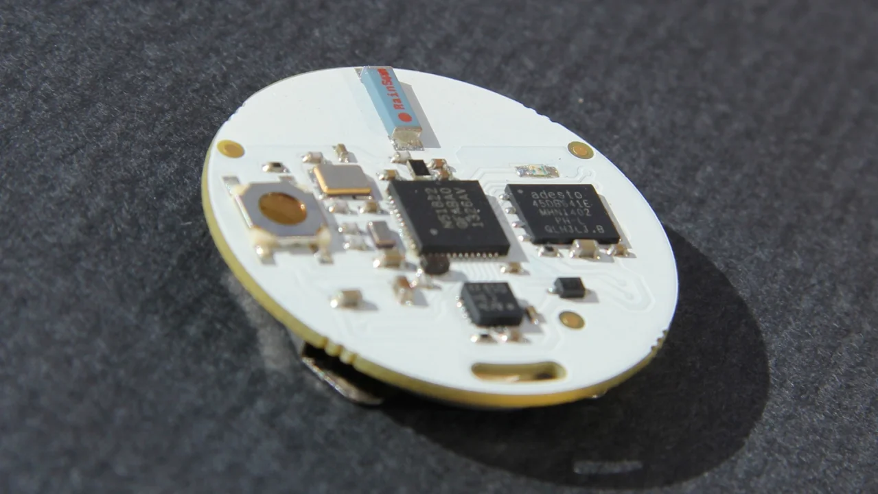 Close-up of the round white OpenBeacon proximity tag circuit board lying on a grey surface, showing the radio chip, flash memory, accelerometer and chip antenna