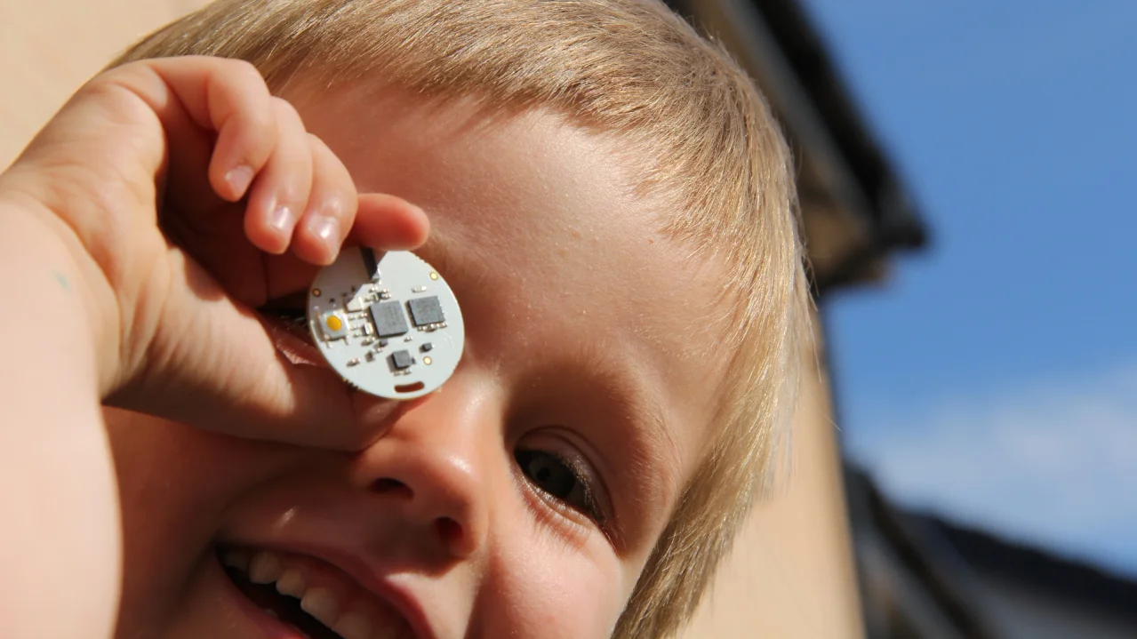 A smiling child holding the small OpenBeacon tag board up to the camera