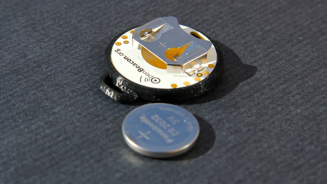 OpenBeacon tag in its round black 3D-printed housing, opened to show the board, next to a CR2032 coin cell
